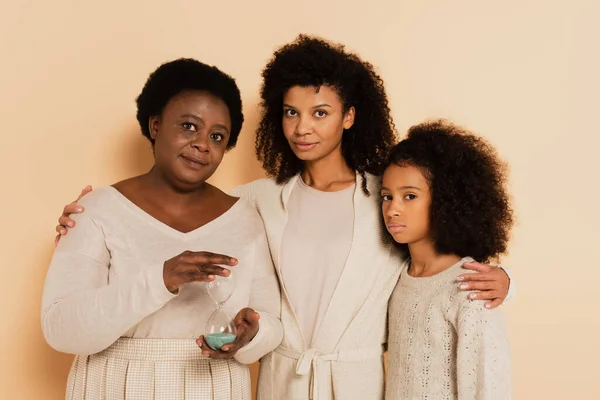 深刻なアフリカ系アメリカ人の祖母は砂時計を持っていて娘と孫娘はベージュの背景に立っています — ストック写真