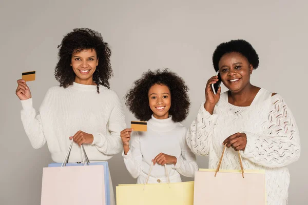 african american mother and daughter holding credit cards and shopping bags near grandmother speaking on phone isolated on grey