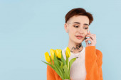 fiatal tetovált nő narancs kardigán gazdaság tulipán, miközben beszél okostelefon elszigetelt kék