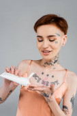 boldog fiatal nő tetoválás tartó cső és alkalmazása testápoló, miközben mosolyogva elszigetelt szürke 