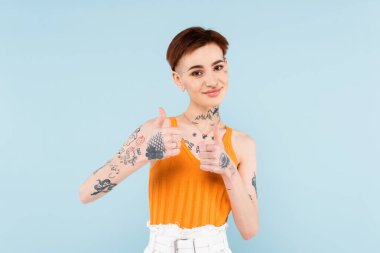 pleased and tattooed woman showing thumbs up isolated on blue  clipart