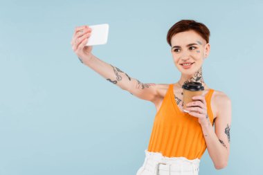 Gülümseyen ve dövmeli bir kadın akıllı telefondan selfie çekerken elinde kahve tutarken mavi ışıkta izole oluyor.