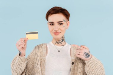 Neşeli ve dövmeli bir kadın, maviyle izole edilmiş kredi kartını işaret ediyor.