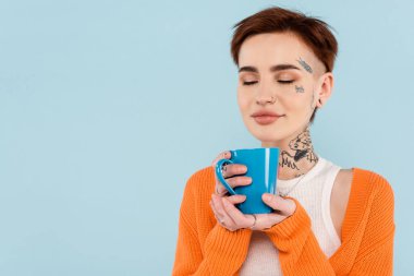 Mavi kahve koklarken elinde turuncu hırka tutan mutlu ve dövmeli bir kadın.