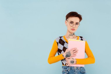 Gözlüklü genç öğrenci kameraya bakıyor ve mavi klasörü tutuyor.