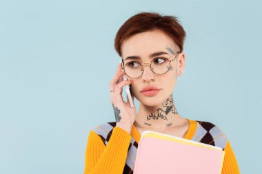 Gözlüklü genç bir kadın cep telefonuyla konuşuyor ve mavi klasörü tutuyor.