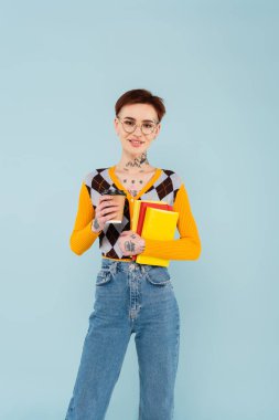 Mutlu dövmeli gözlüklü bir öğrenci elinde kitap ve kağıt bardakla maviden izole edilmiş.