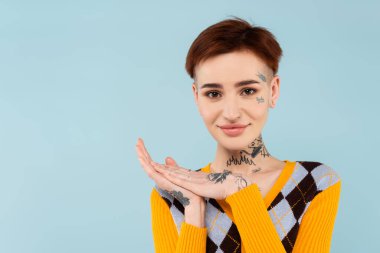 Gülümseyen genç ve dövmeli bir kadın mavi üzerine izole edilmiş kameraya bakıyor.