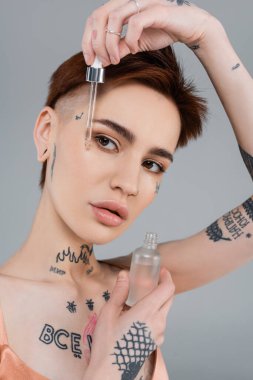 young woman with tattoos holding bottle with serum and pipette isolated on grey  clipart