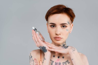 tattooed young woman holding bottle with serum isolated on grey  clipart