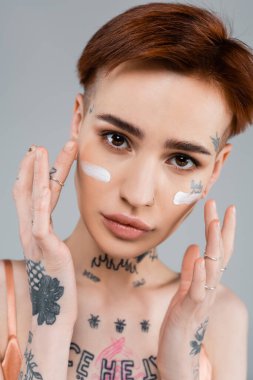 Dövmeli kadın griye izole edilmiş nemlendirici yüz kremi sürüyor. 