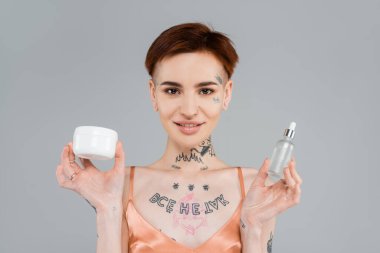 young and tattooed woman holding cosmetic products while smiling isolated on grey  clipart