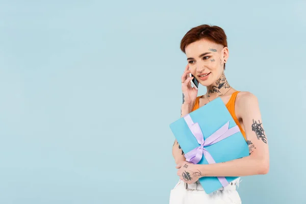 快乐的 纹身的女人拿着包裹着的礼物 一边用蓝色隔离的手机交谈 — 图库照片