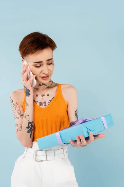 Young Tattooed Woman Holding Wrapped Present Talking Cellphone Isolated Blue — Stock Photo, Image