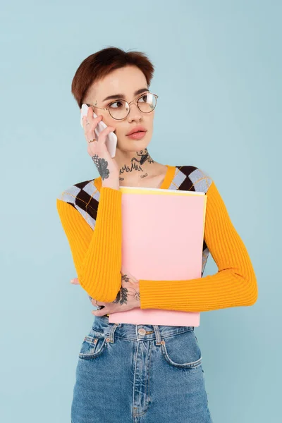 Jovem Estudante Tatuado Óculos Falando Celular Segurando Pasta Isolada Azul — Fotografia de Stock