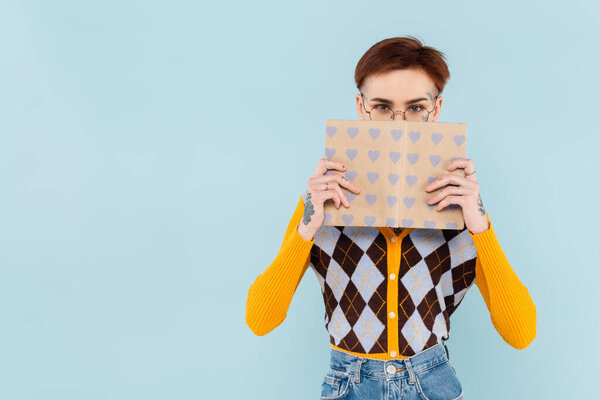 young tattooed student in glasses covering face with book in heart pattern cover isolated on blue