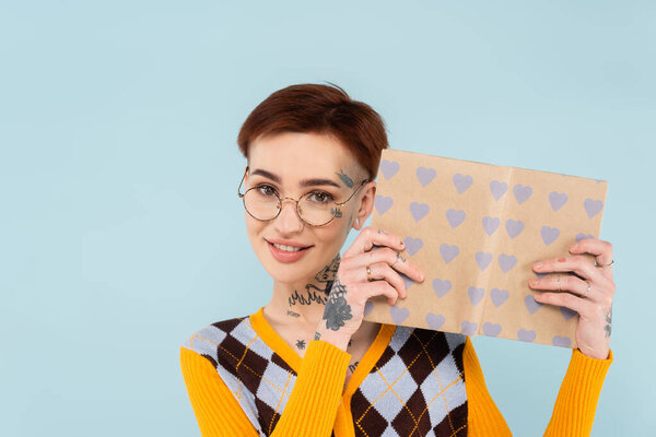 happy tattooed woman in glasses holding book in heart pattern cover isolated on blue