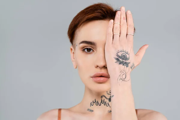 young tattooed woman looking at camera and covering face with hand isolated on grey