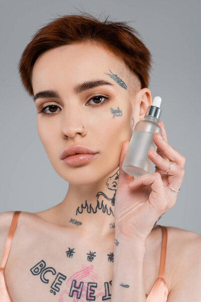 татуированная молодая женщина держит бутылку с увлажняющей сывороткой, изолированной на сером 