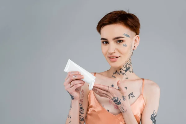 tattooed young woman holding tube and applying body lotion while smiling isolated on grey