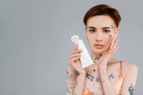 young woman with tattoos holding tube and applying face cream isolated on grey