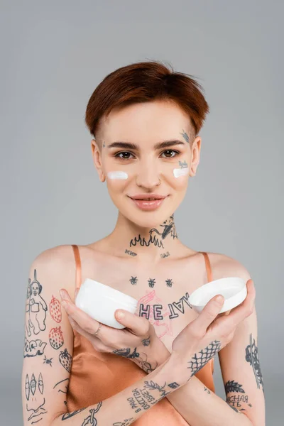 tattooed and happy woman with cosmetic cream on face holding container isolated on grey
