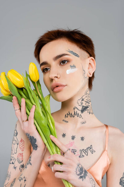 Татуированная молодая женщина с косметическим кремом на лице, держащая тюльпаны изолированные на сером 