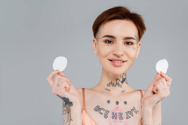 happy and tattooed woman holding cotton pads isolated on grey 