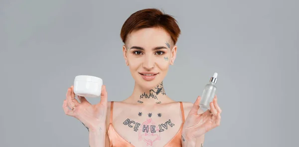 Mujer Joven Tatuada Sosteniendo Productos Cosméticos Mientras Sonríe Aislada Gris — Foto de Stock