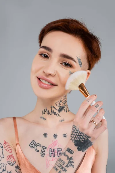 明るい入れ墨の女性は灰色に隔離された顔の粉を適用しながら 化粧ブラシを保持 — ストック写真