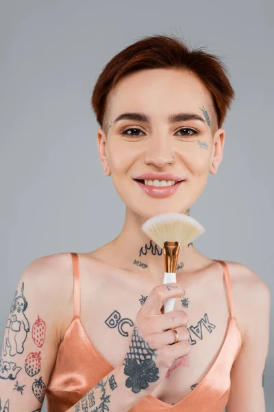 happy tattooed woman holding cosmetic brush isolated on grey