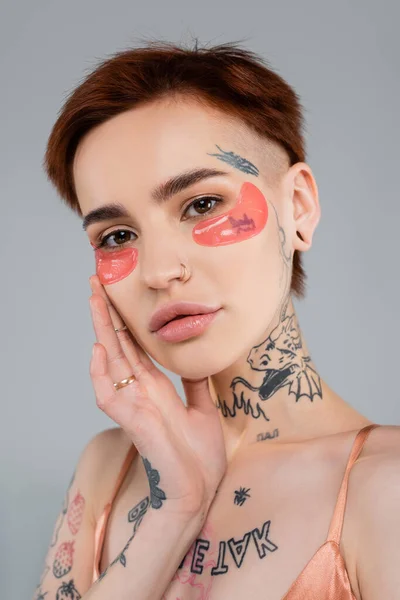 young and tattooed woman with moisturizing eye patches isolated on grey