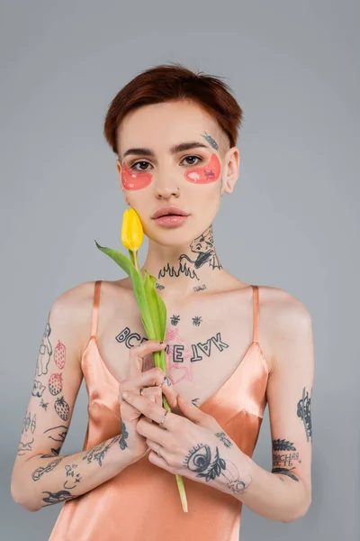 young and tattooed woman in pink eye patches holding tulip isolated on grey