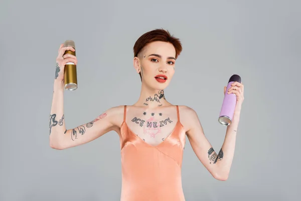 young tattooed woman with red lips holding hairspray bottles isolated on grey