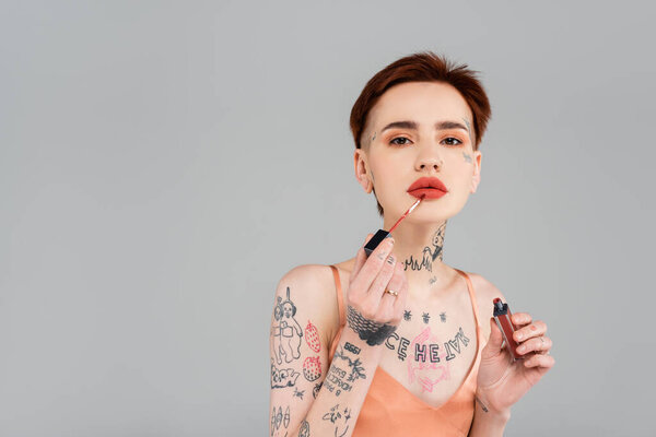 молодая татуированная женщина с красными губами наносит блеск для губ, изолированный на сером