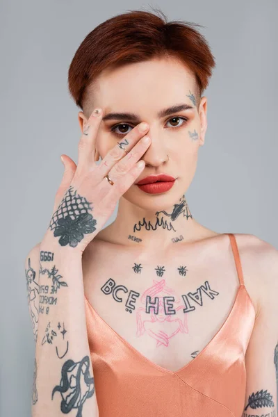 tattooed young woman with red lips covering face with hand isolated on grey