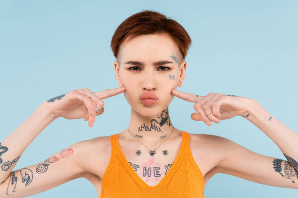 woman with tattoos and orange knitted tank top pointing at puffing cheeks isolated on blue