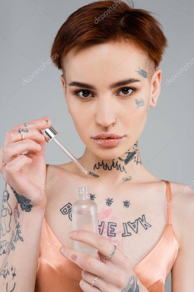 Young tattooed woman holding bottle with serum and pipette isolated on grey