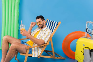 Smiling man with orange juice sitting on deck chair near suitcase, swimming flippers and inflatable ring on blue background  clipart