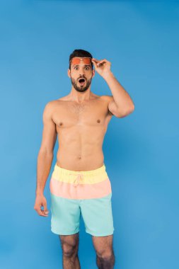 Mavide güneş gözlüğü tutan şaşkın, üstsüz adam. 