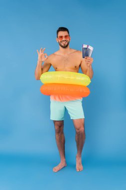 Smiling man in inflatable rings showing ok gesture and holding passports on blue background  clipart