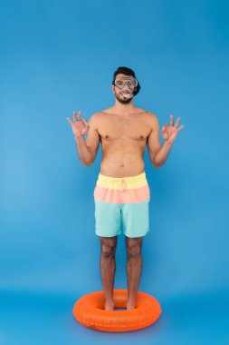 Shirtless man in swimming goggles showing ok gesture while standing in inflatable ring on blue background  clipart