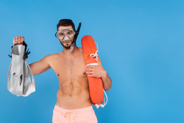 Shirtless man in swimming goggles holding life buoy and flippers isolated on blue 