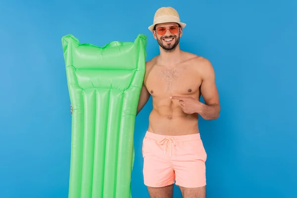 戴太阳镜的无袖男人指着蓝色隔离的充气床垫 — 图库照片