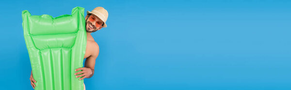 Веселый человек в солнцезащитных очках и шляпе с надувным матрасом, изолированным на голубом 
