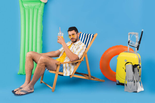Man in sunglasses holding orange juice on deck chair near inflatable ring and suitcase on blue background 