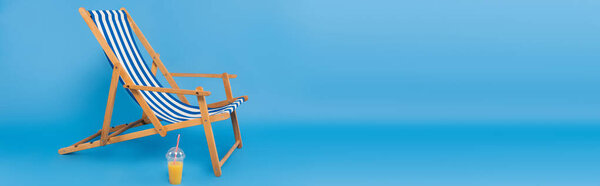 Orange juice in plastic cup near deck chair on blue background, banner 