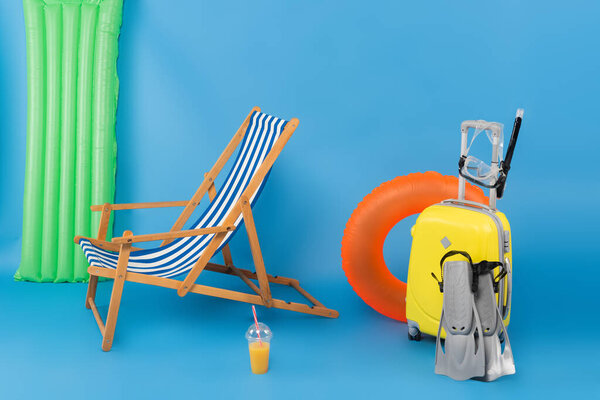 Deck chair near suitcase, swimming goggles and orange juice on blue background 