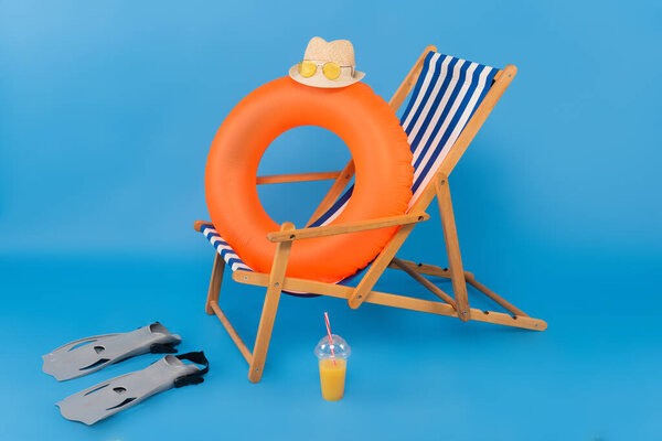 Straw hat on inflatable ring near swimming flippers and orange juice on blue background 