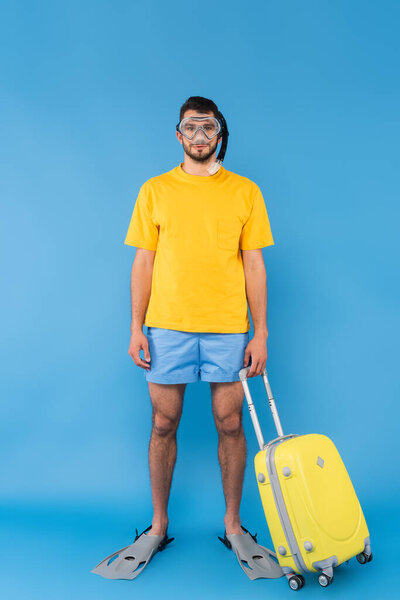 Young man in swimming goggles and flippers holding suitcase on blue background 